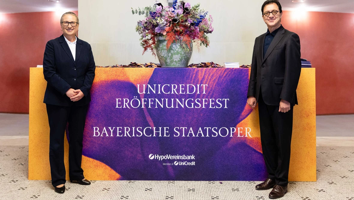 Zwei Personen stehen neben einer Theke mit Blumen und dem Schriftzug Unicredit Eroeffnungsfest Bayerische Staatsoper.