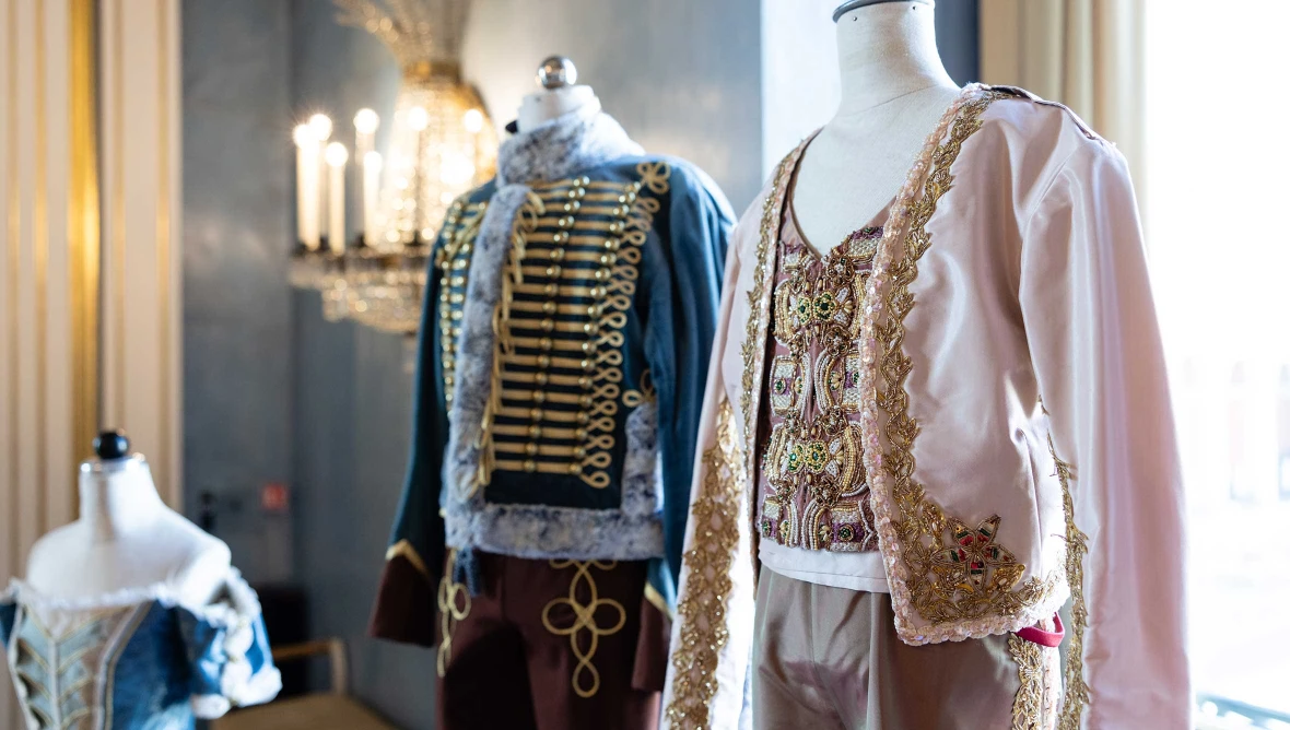 Historische Kostueme auf Schneiderpuppen in einem Innenraum mit Fenstern und dekorativer Beleuchtung.