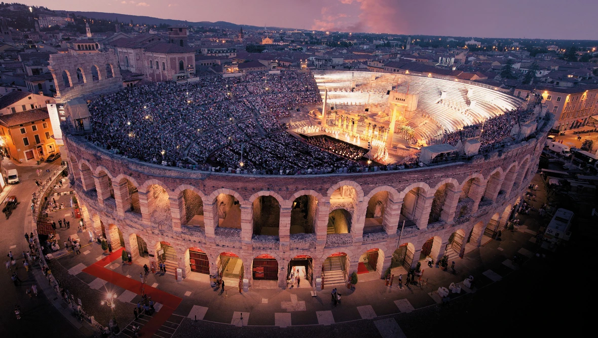 Aufnahme eines römischen Amphitheaters von oben bei Nacht, die Zuschauerränge sind voll besetzt und ganz vorne ist eine beleuchtete Bühne zu sehen. 