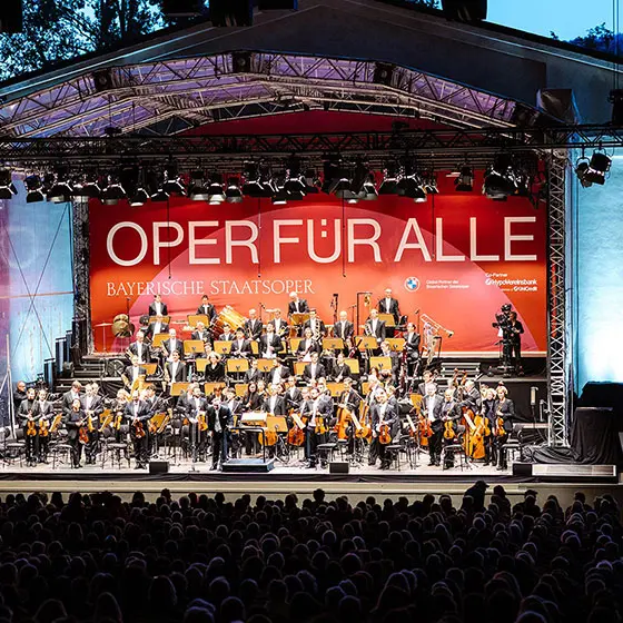 Konzertbühne mit Orchester, am Bühnenhintergrund ist ein großes rotes Plakat mit der Aufschrift "Oper für Alle".