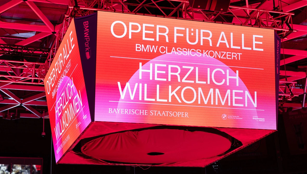 Großer Bildschirm zeigt ein Motiv mit rot-gemustertem Hintergrund und der Überschrift Herzlich Willkommen und  "Oper für Alle"