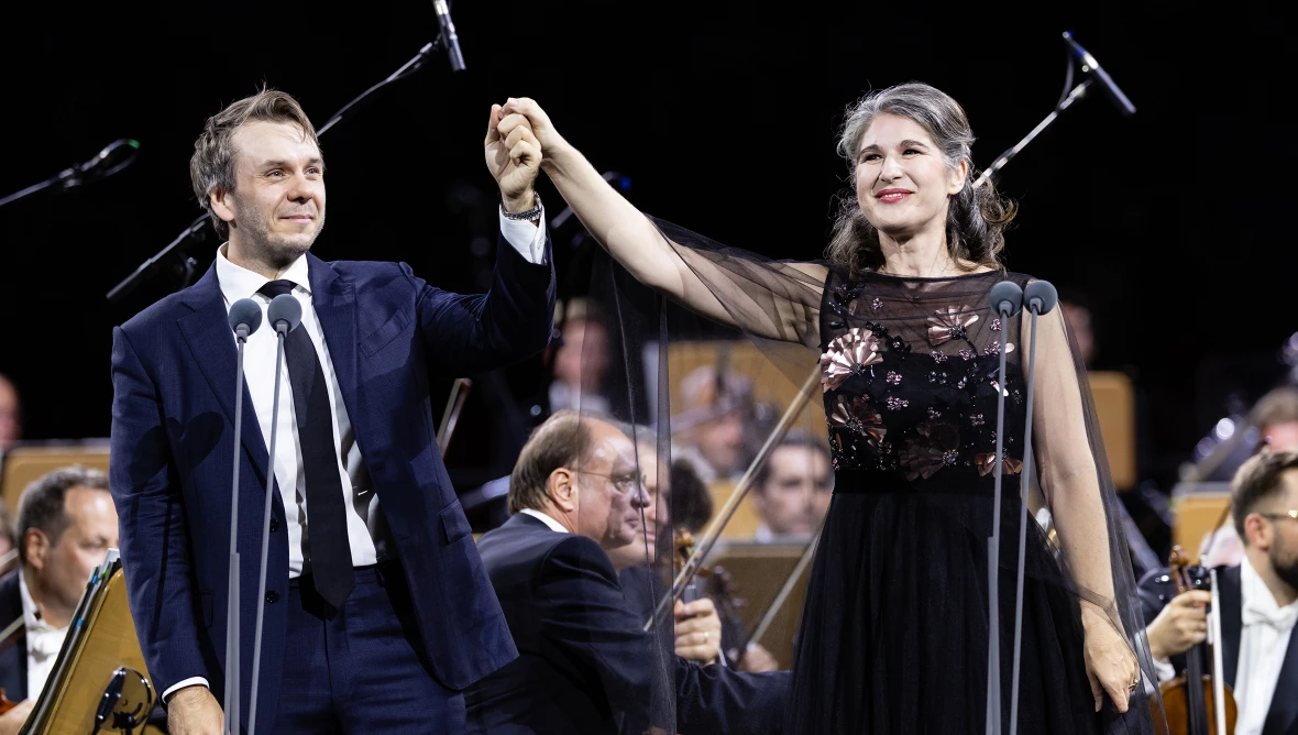 Zwei Solist:innen, ein Herr im Anzug und eine Dame im schwarzen Abendkleid stehen vor einem Orchester auf der Bühne und halten zum Applaus die Hände erhoben.
