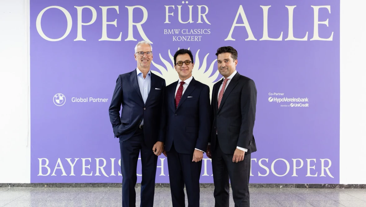 Drei Herren in dunklen Anzügen stehen vor einem großen lilafarbenem Plakat auf dem in gelber Schrift Oper für alle steht.