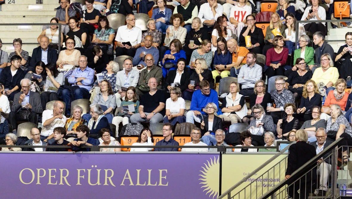 Blick auf die Zuschauerränge im Detail, unterschiedliche Personen sind auf den Sitzplätzen zu sehen, unten ist ein Banner mit der Aufschrift Oper für alle.