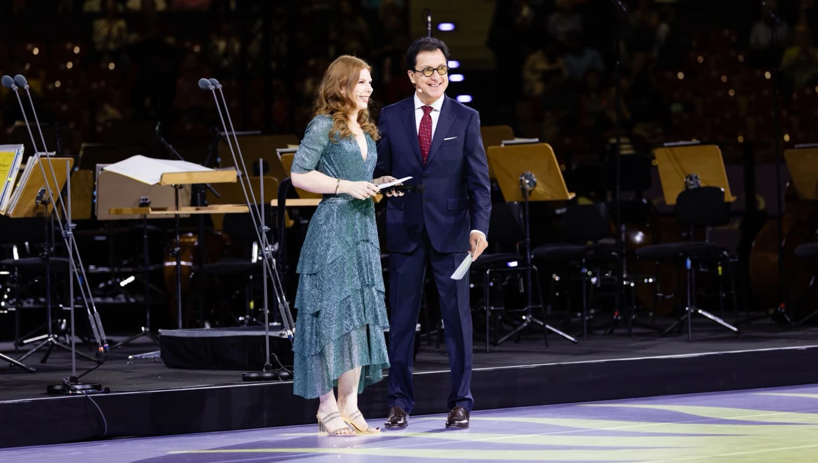 Eine Frau im türkisen Abendkleid und ein Herr in dunklem Anzug stehen vor unbesetzten Plätzen eines Orchesters im Hintergrund.