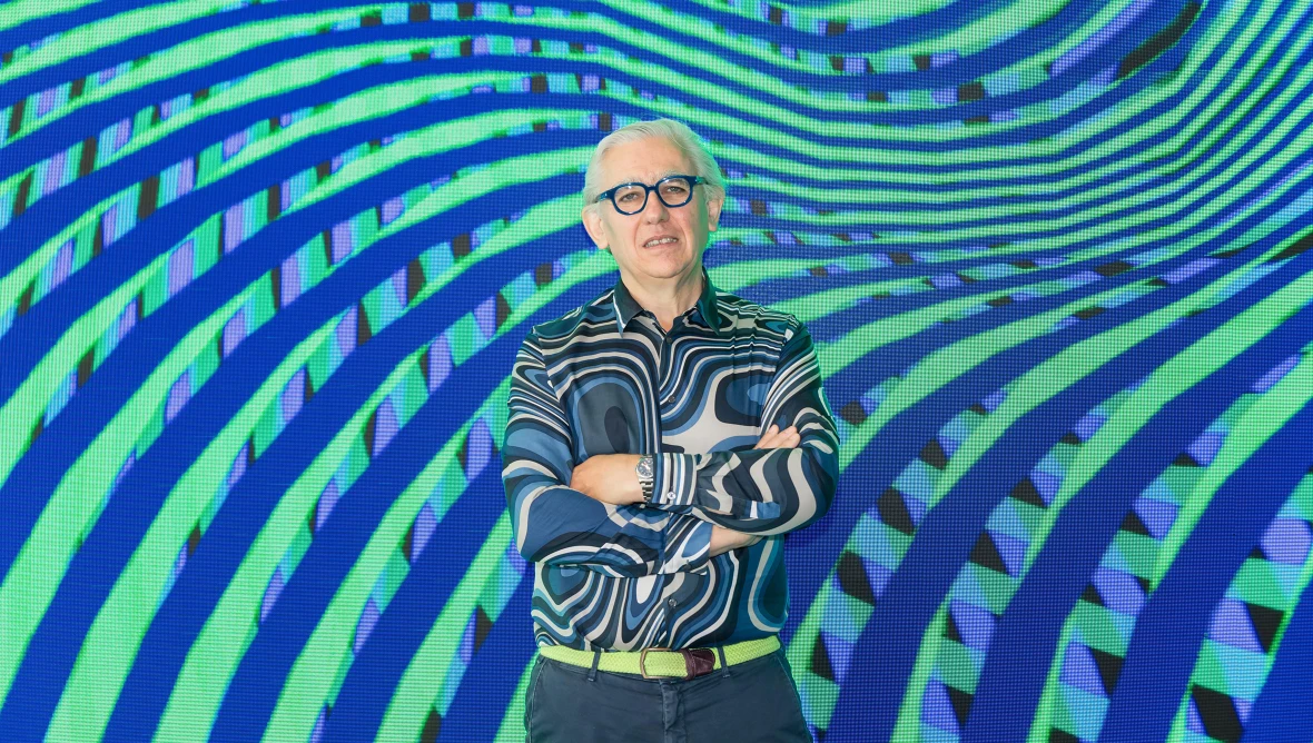 Der Künstler Miguel Chevalier, ein älterer Herr mit weißen Haaren, einer schwarzen Brille und einem bunt-gemustertem Hemd steht vor einem bunten Hintergrund. Dieser zeigt wellenartige Formen in blau- und grün-Tönen.
