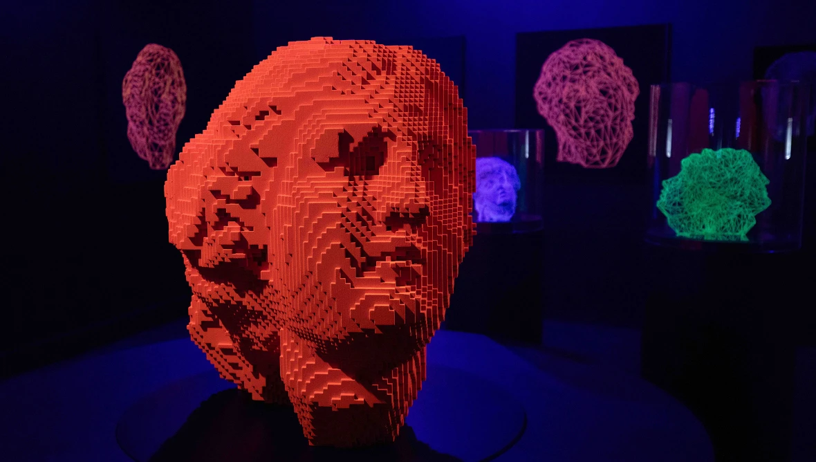 Eine leuchtend orangefarbene, pixelartig strukturierte, kugelförmige Skulptur vor dunklem Hintergrund, die wie ein futuristisches fragementiertes Gesicht wirkt. 