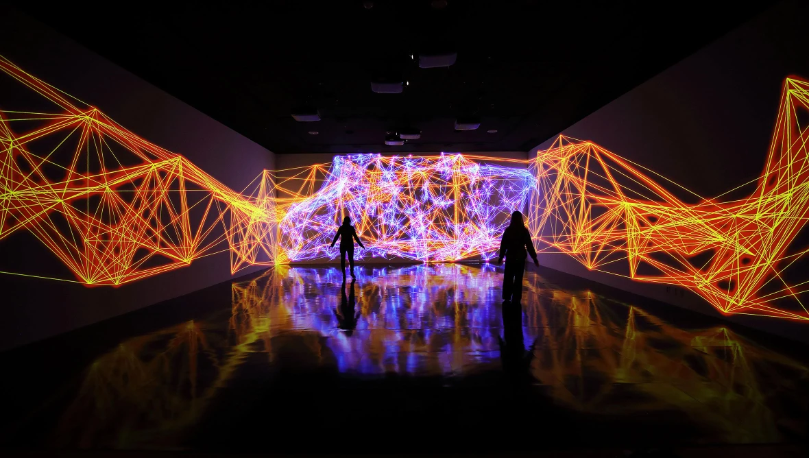 Im dunklen Raum füllen leuchtende, netzartige Strukturen in orange und violett die Wände - eine digitale, raumgreifende Landschaft, in der Silhouetten von Besucher:innen zu erkennen sind. 