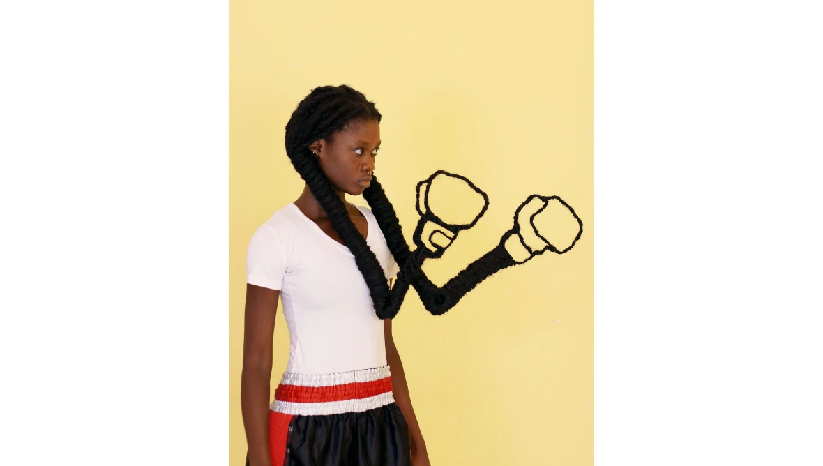 Ein Foto eines jungen Mädchens mit einer Sporthose und einem weißen T-Shirt, ihre langen schwarzen Haare sind zu Zöpfen geflochten an deren Enden sich zwei Boxhandschuhe aus Haaren formieren. Der Hintergrund ist gelb. 