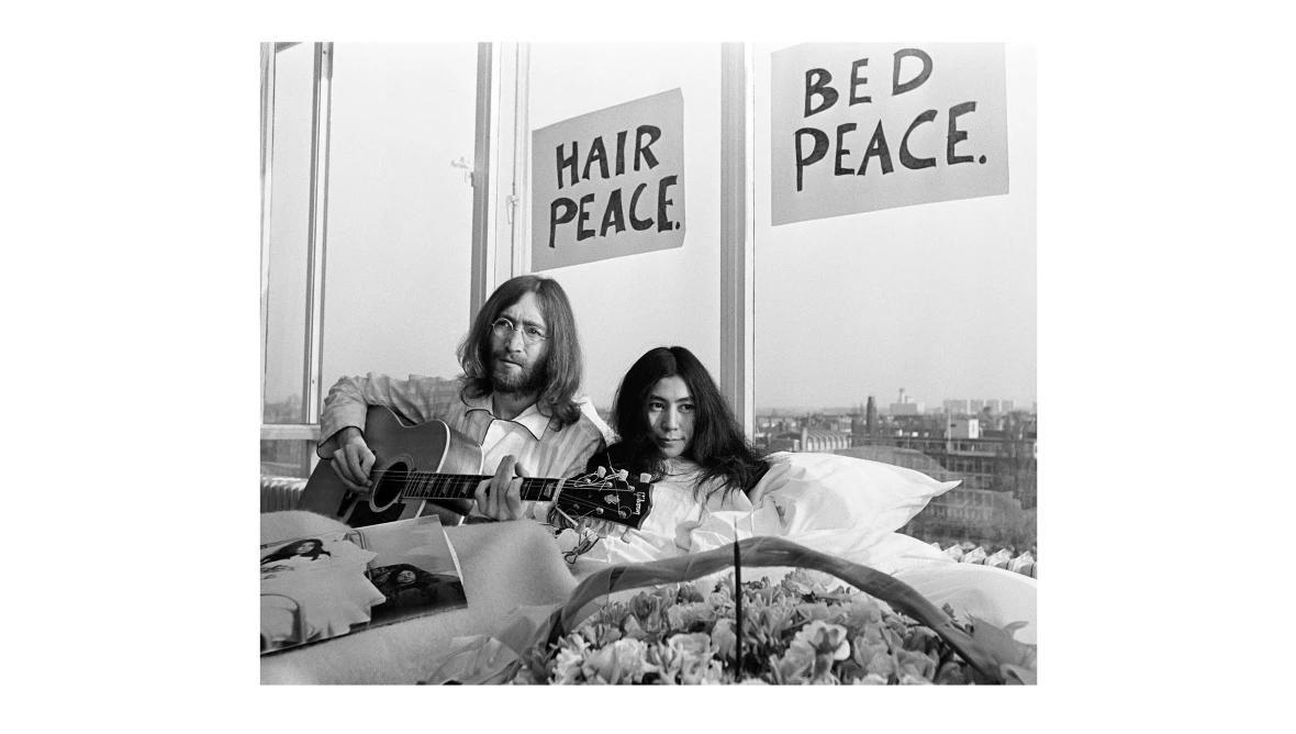 Eine schwarz-weiß Aufnahme von John Lennon (links) und Yoko Ono (rechts). Beide liegen im Bett, John Lennon spielt Gitarre und im Hintergrund sind zwei Plakate mit der Aufschrift "Hair Peace" und "Bed Peace" zu sehen. 