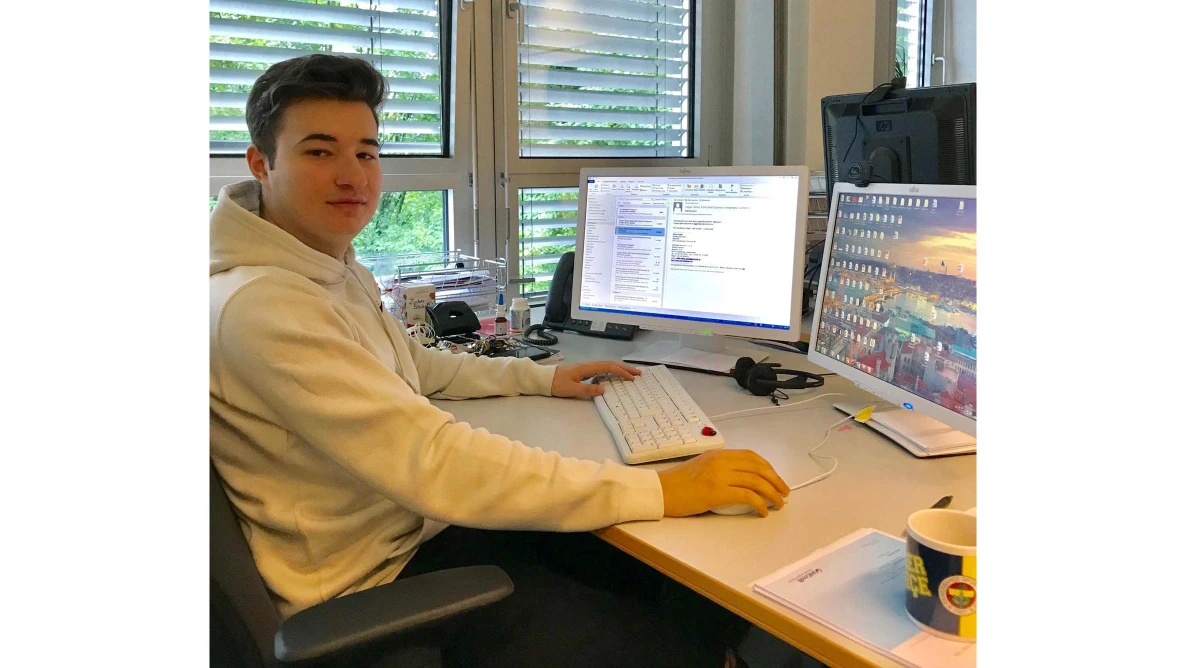 Junge Person sitzt an Schreibtisch mit zwei Monitoren und schaut zur Kamera