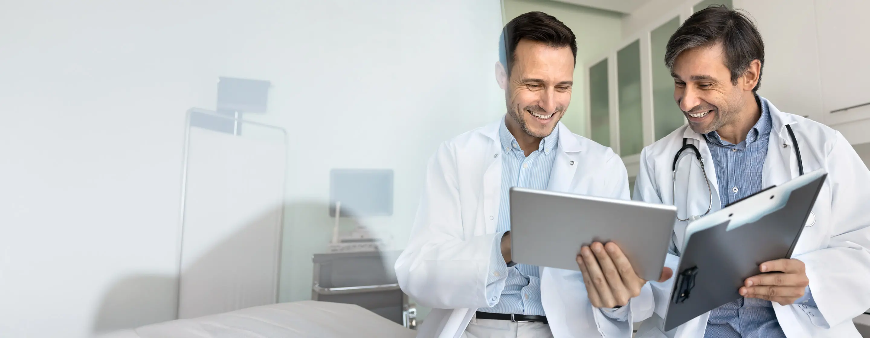 Zwei Ärzte im weißen Kittel schauen gemeinsam lachend auf ein Tablet und eine Patientenakte.