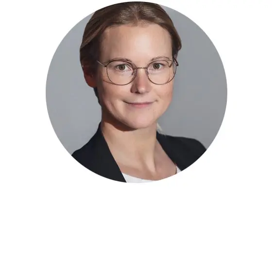Porträt von Dörte Andratschke mit runder Brille und schwarzem Blazer vor neutral grauem Hintergrund.