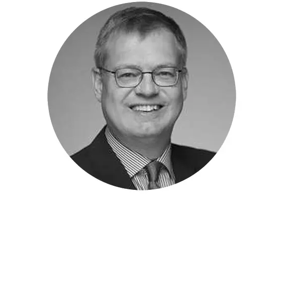 Eine Person in einem formellen Anzug und Brille lächelt und schaut in die Kamera. Das Bild ist schwarz weiß.