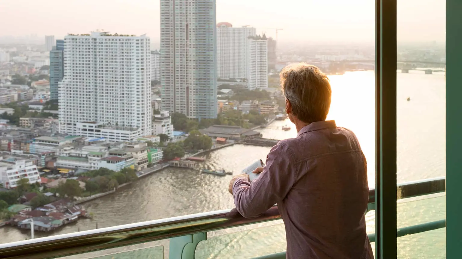 Person mit grauen Haaren steht auf einem Balkon und blickt bei Sonnenuntergang auf einen Fluss und eine Stadt mit Hochhäusern.