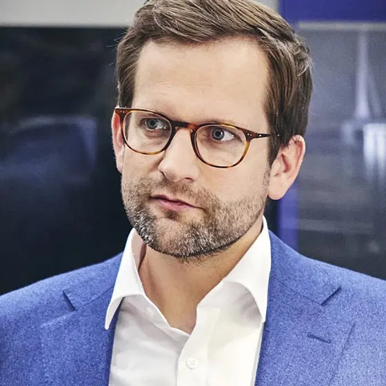 Person mit runder Brille in blauem Sakko und weißem Hemd blickt leicht zur Seite vor unscharfem Innenraumhintergrund in kühlen Farben.