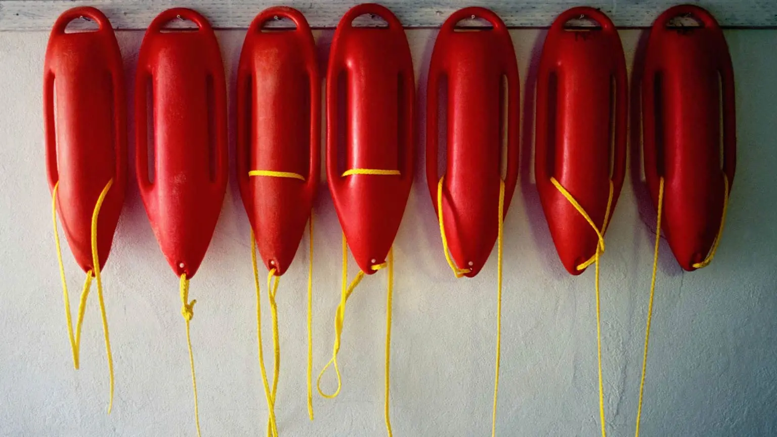 Acht rote Rettungsbojen mit gelben Seilen hängen gleichmäßig an einer hellen Wand nebeneinander.