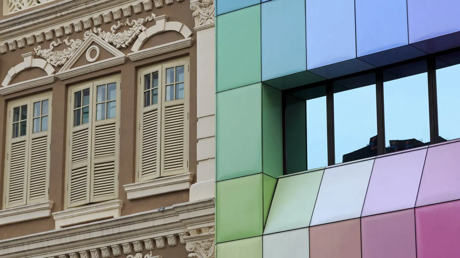 Klassische Fassade mit Ornamenten und Fensterläden neben moderner Fassade mit farbigen Glasflächen.