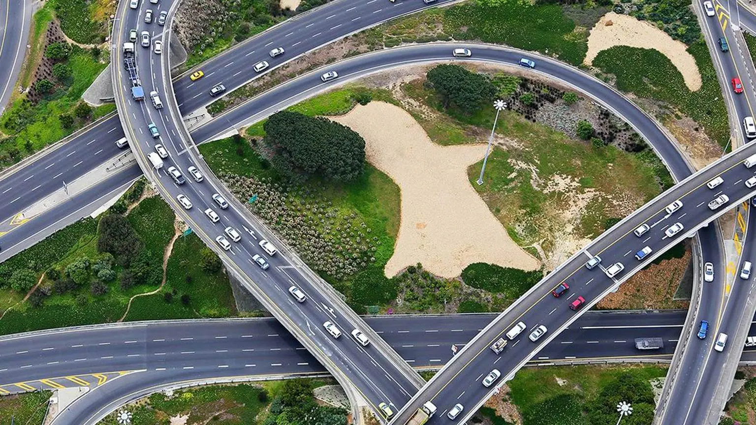 "Luftaufnahme eines Autobahnkreuzes mit mehreren Ebenen und vielen Fahrzeugen auf verschiedenen Fahrspuren zwischen Grünflächen. "