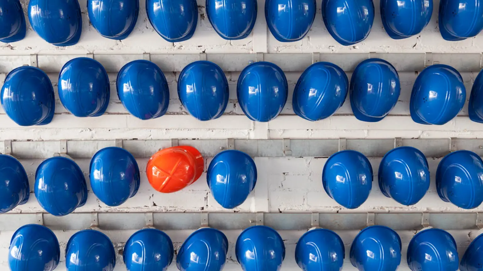 Viele blaue Schutzhelme hängen an einer Wand, dazwischen ein auffälliger orangefarbener Helm.