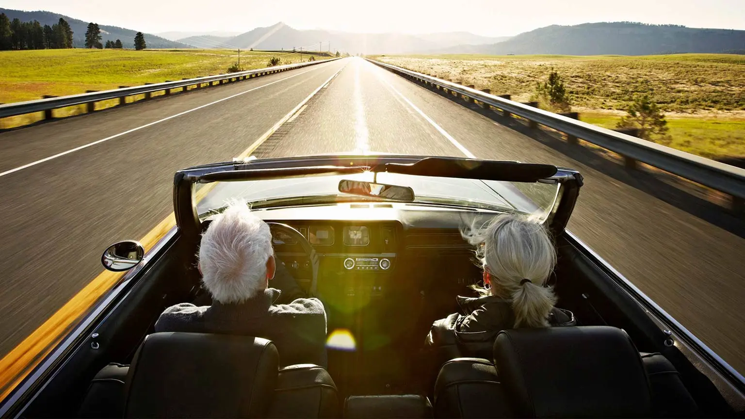 Zwei ältere Personen fahren in einem Cabrio über eine sonnige Landstraße mit Feldern und Hügeln.