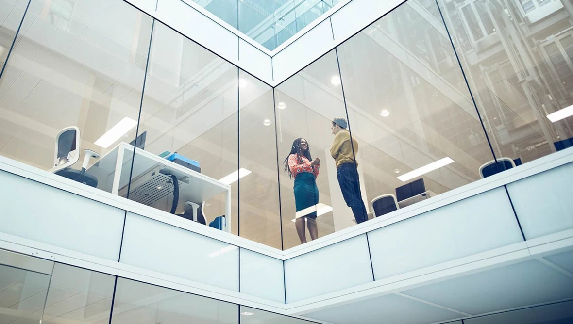 Zwei Personen stehen in einem modernen Bürogebäude hinter großen Glasfassaden und sprechen miteinander.