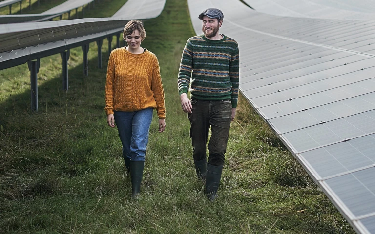 Zwei Personen gehen zwischen Solarpaneelen über eine Wiese, beide tragen Pullover, Gummistiefel und blicken leicht nach unten.