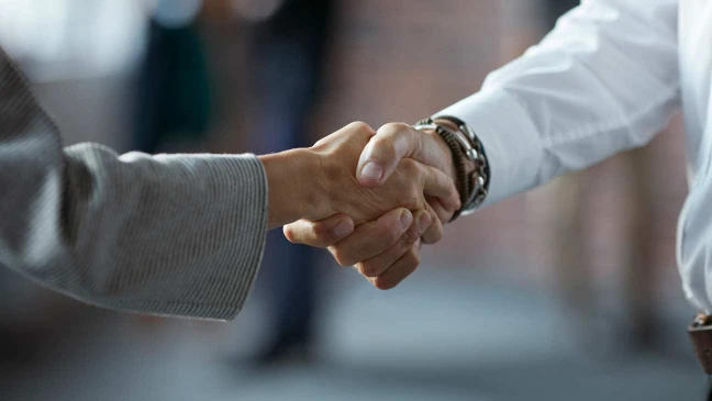 Zwei Personen geben sich die Hand, sichtbar sind nur die Hände im Vordergrund, der Hintergrund ist unscharf.