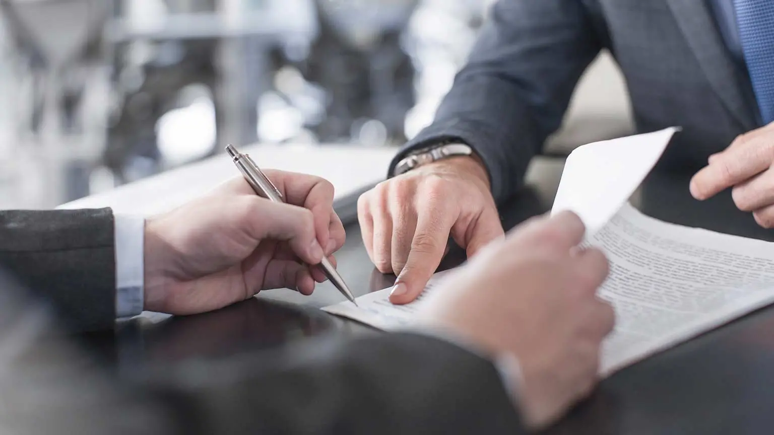 Zwei Personen in Anzügen besprechen ein Dokument am Tisch, eine Person zeigt, die andere hält einen Stift.