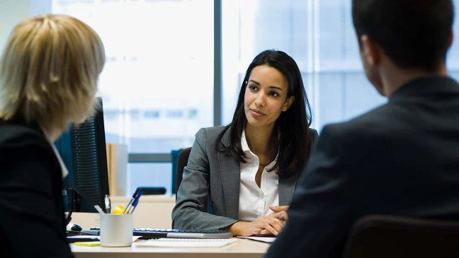 Drei Personen im Businessumfeld sitzen an einem Tisch und führen ein Gespräch in einem modernen Büro.