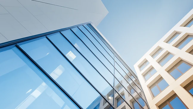 Moderne Gebäudeansichten aus leichtem Aufwärtswinkel: links eine Fassade aus Glas und Stahl, rechts ein helles Gebäude mit geometrischen Fensterrahmen, vor klarem blauem Himmel.