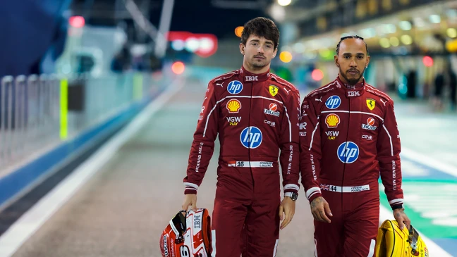 Ferrari-Fahrer in Rot: Lewis Hamilton und Charles Leclerc posieren mit Helmen und Sponsorenlogos vor einfarbig rotem Hintergrund.