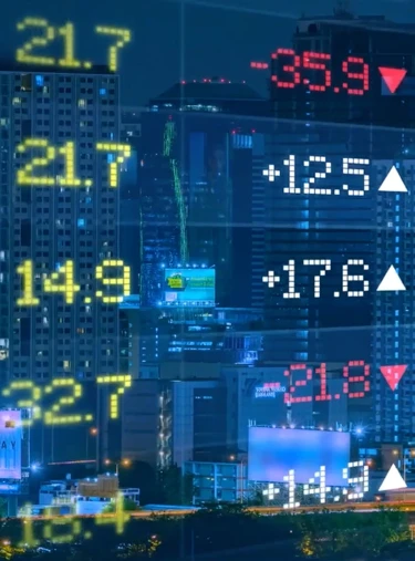 Digitale Börsenanzeige mit Zahlen und Pfeilen in verschiedenen Farben vor nächtlicher Stadtkulisse mit beleuchteten Hochhäusern.