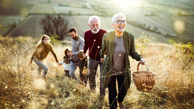 Gruppe von fünf Personen geht gemeinsam durch eine hügelige Landschaft mit trockenem Gras. Eine Person im Vordergrund trägt eine Brille und hält einen Korb, dahinter folgen weitere Personen, darunter auch zwei Kinder. Im Hintergrund sind Felder und sanfte Hügel zu sehen.