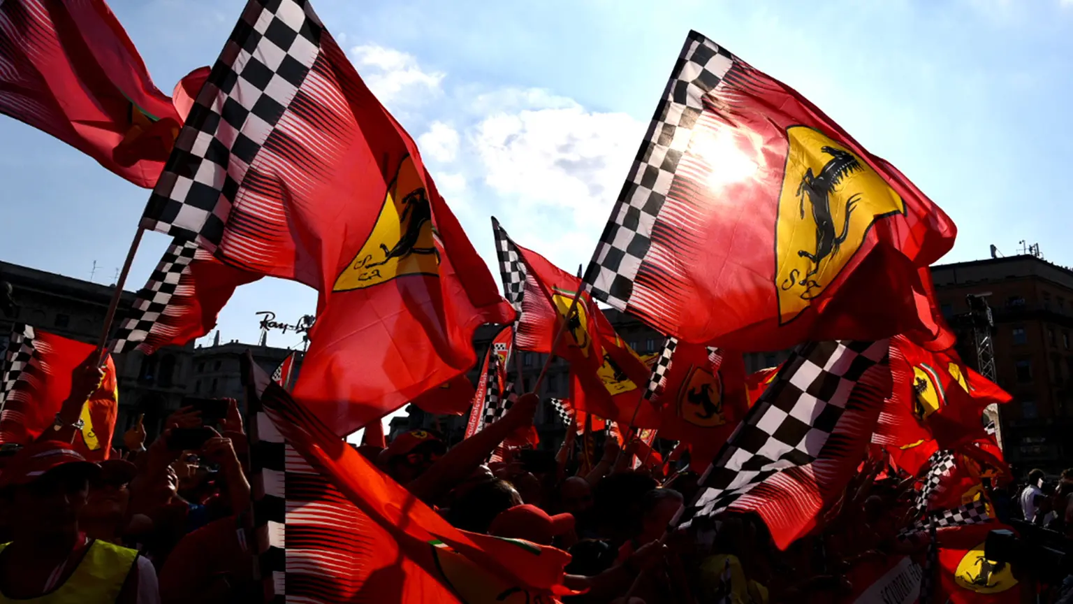 Mehrere rote Ferrari-Flaggen mit schwarzem Pferde-Logo und schwarz-weiß kariertem Rand wehen im Wind vor blauem Himmel.