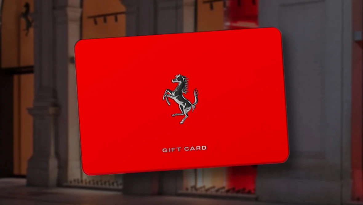Rote Ferrari Gift Card mit schwarzem Ferrari‑Logo vor einer Architekturkulisse mit Säulen und warmem Licht.
