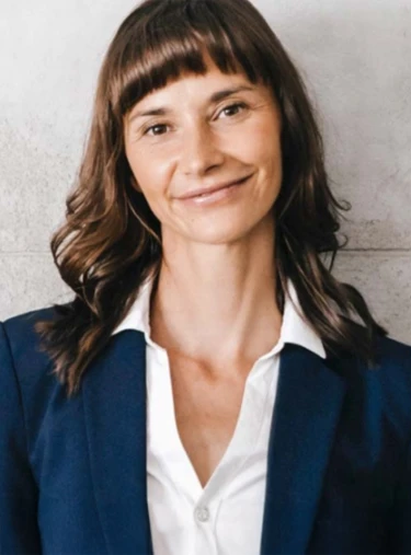 Person in dunkelblauem Sakko und weißem Hemd mit offenem Kragen vor hellgrauer Wand ohne sichtbare Objekte.