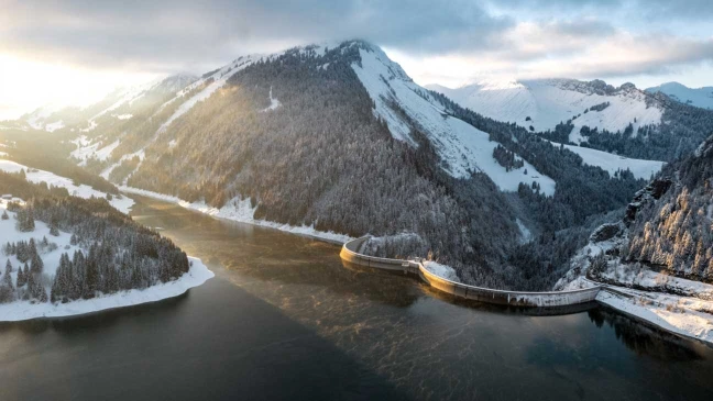 Verschneites Bergtal mit Staumauer über einem Fluss, umgeben von Nadelwald und beleuchtetem Hang unter bewölktem Himmel.