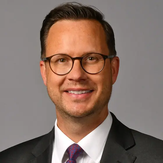 Portrait von Christian Stockfleth. Er trägt einen Anzug mit weißem Hemd und violetter Krawatte, lächelt vor grauem Hintergrund und blickt direkt in die Kamera.